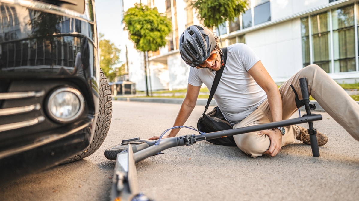 Verkehrsunfall mit E-Scooter, junger Mann mit Knieverletzung, sitzt neben seinem verunfallten Zweirad