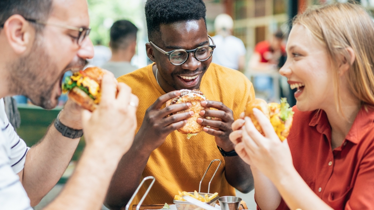 Drei junge Erwachsene an einem Tisch beim Verspeisen von Burgern und Pommes