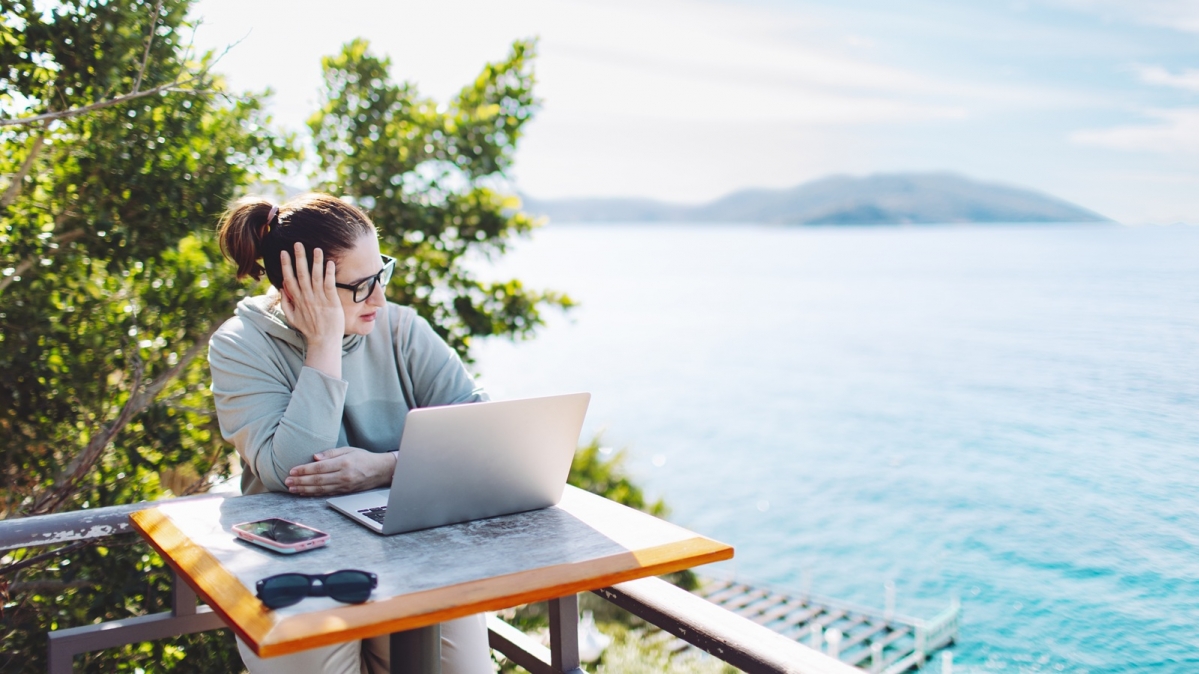 Gefrustete Frau an einem Laptop vor einer schönen Mittelmeerküstenlandschaft