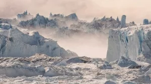 Gletscher mit zerbrochenem Eis, schneebedeckten Bergen im Hintergrund und Nebel über dem Eis.