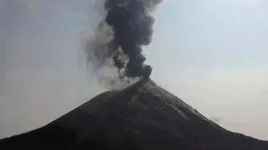 Vulkan spuckt schwarzen Rauch in den Himmel, Wasser im Vordergrund, Sonne im rechten oberen Bereich.