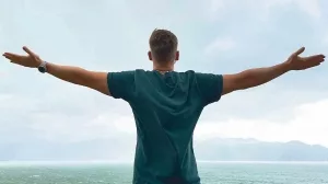 Junger Mann mit ausgestreckten Armen blickt auf das Meer und den Himmel.