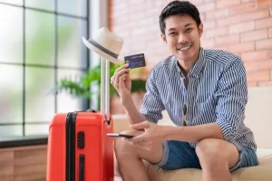 Junger Mann mit gepacktem Koffer und Smartphone und Kreditkarte in den Händen
