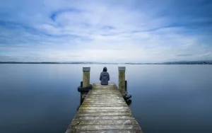 Einsamkeit; junge Frau, die allein auf einem Steg an einem dunklen See sitzt
